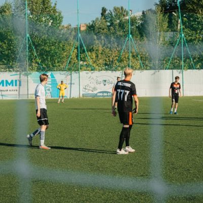 Flera unga människor spelar fotboll på en utomhusbana omgiven av ett staket, med träd och reklamskyltar i bakgrunden.